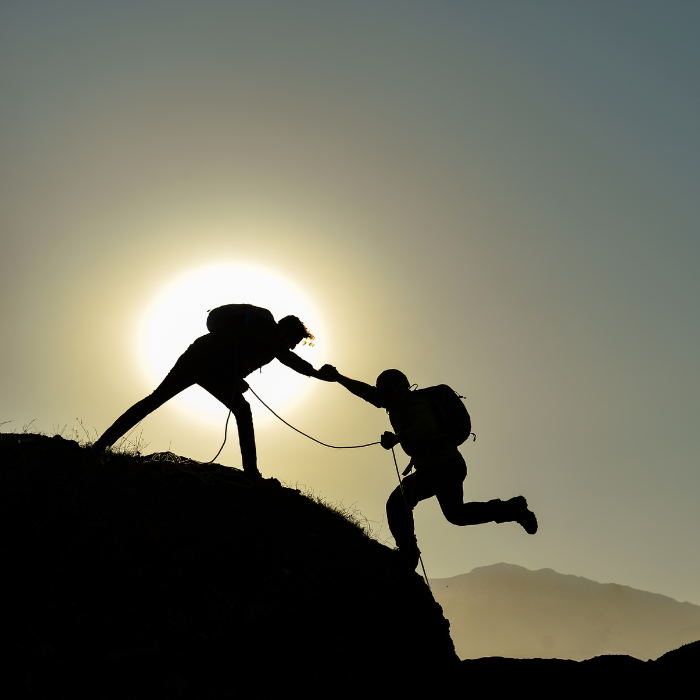 Silhouette of a person standing on a mountain peak pulling another person up.