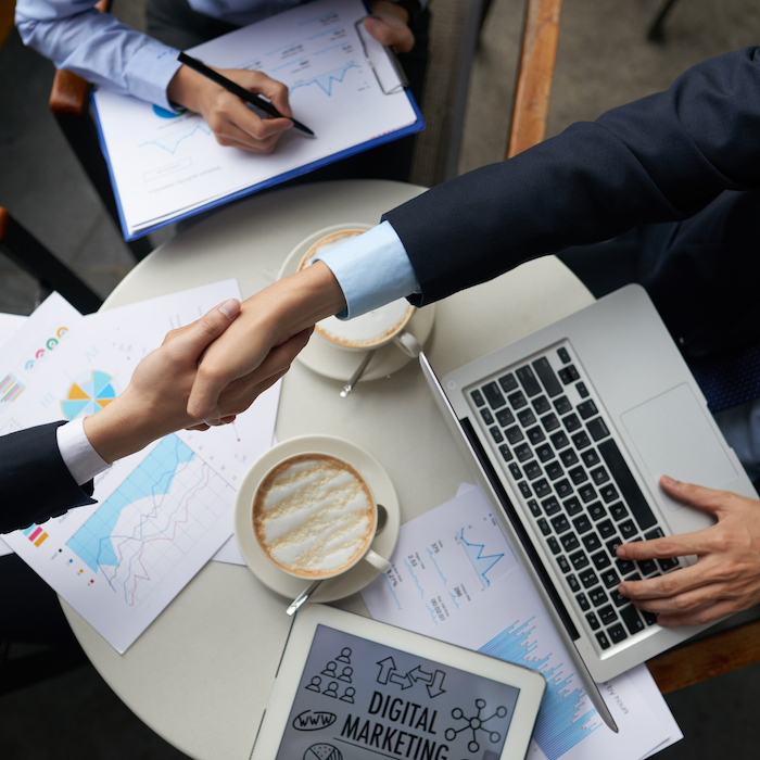 Two people shaking hands over a table with a laptop and charts.